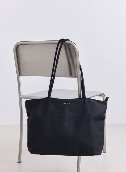 Musta nailoninen tote-laukku valkoisella aim'n-logolla roikkuu minimalistisella metallituolilla.