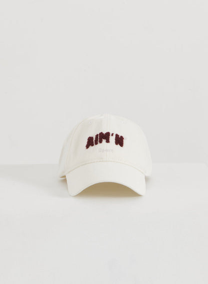 Kermavärinen puuvillainen Campus Cap tummanpunaisella pörröisellä aim'n-logolla minimalistisessa flatlay-kuvassa.