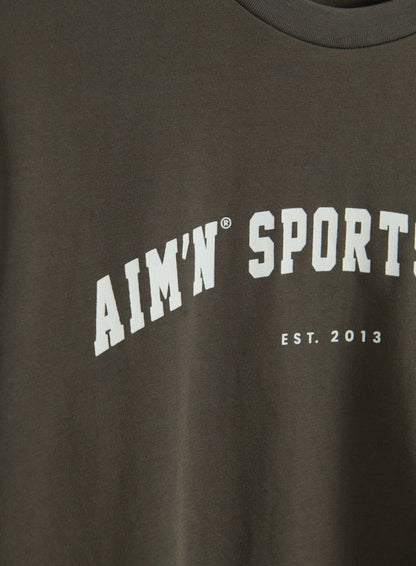 Lähikuva Dark Ivy -värisen Varsity Boxy -T-paidan ribatusta pyöreästä pääntiestä ja valkoisesta aim'n Sport Est. 2013 -grafiikasta