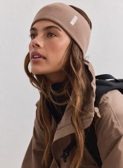 Hasselpähkinänvärinen ribbineulottu Outdoor Logo Headband valkoisella aim'n-logolla, beigen takin ja repun kera.
